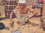  Mwadana crouches before a charcoal fire and thrusts the iron spike into the coals. When it is red hot, he scrapes the impurities from it.
