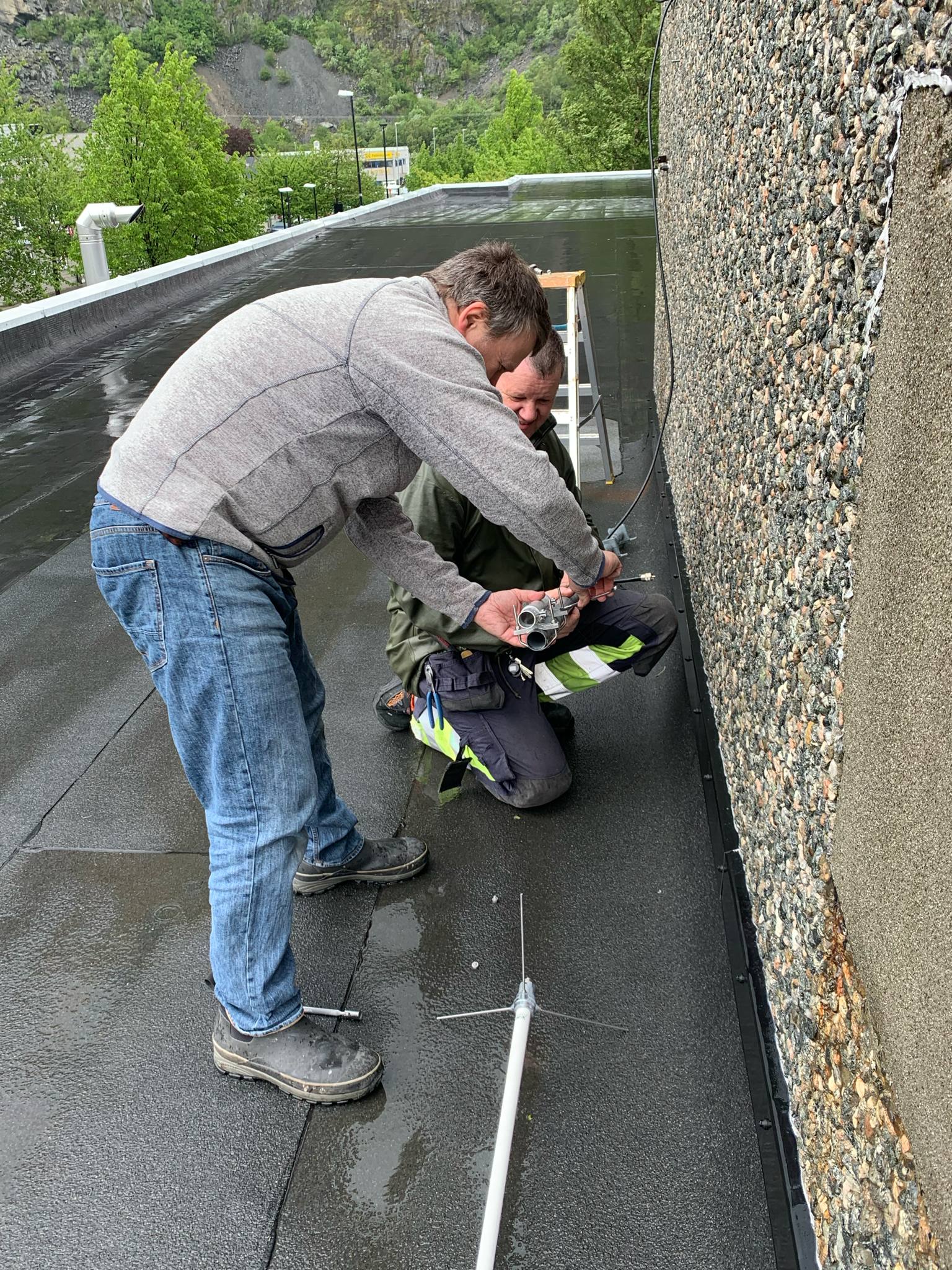 To radioamatører arbeider med antennemontering på rådhustaket i Sunndalsøra, knelende ved bygningsveggen med antennedeler på bakken