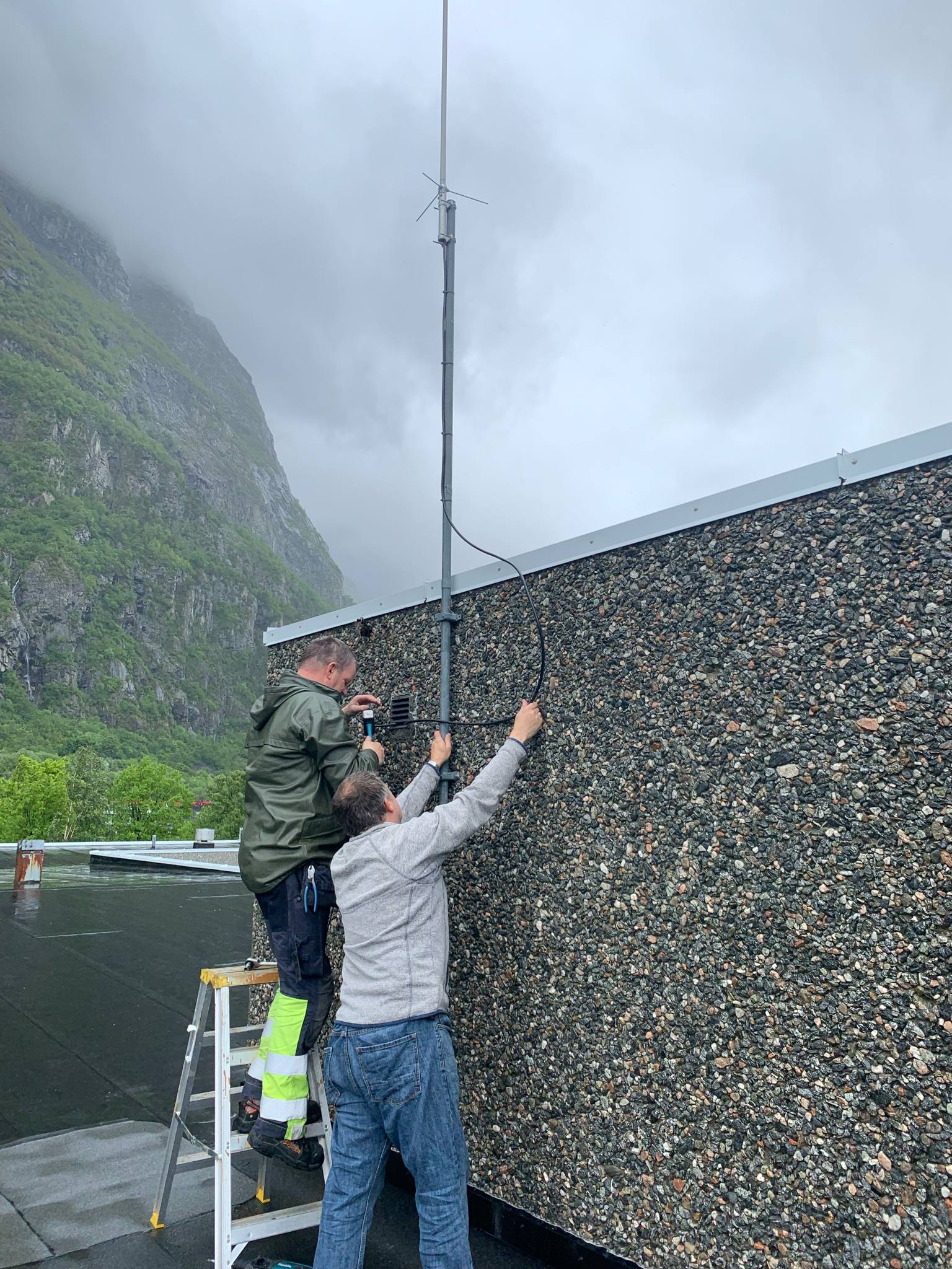 To radioamatører monterer antenne på veggen av rådhuset, den ene står på stige, med fjell og skyer i bakgrunnen