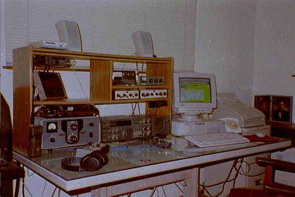 Picture of the ham shack with the Kenwood TS-940 on the desk with the Collins 30L1, and the computer.  Accessories are on shelving above the HF rig on the desk.  An old Model 15 teletype machine is almost out of sight to the left of the desk.